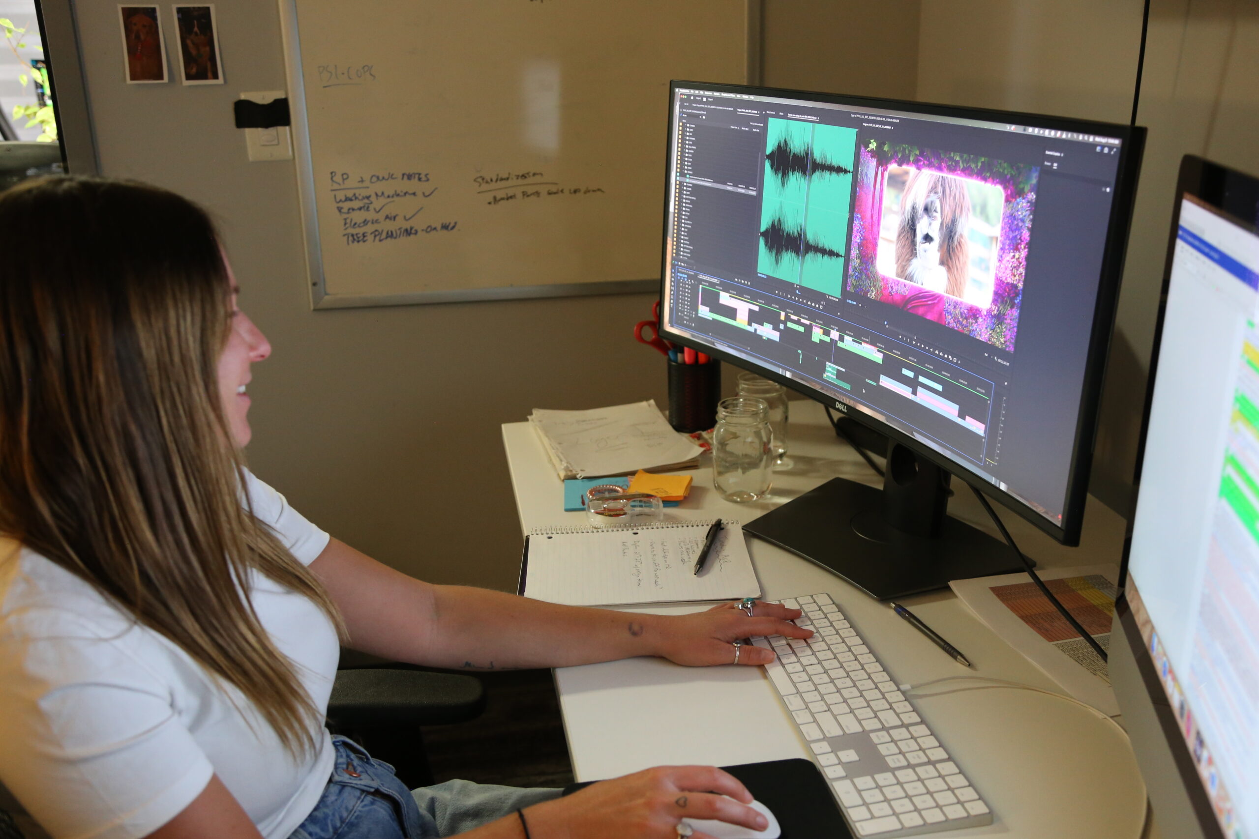 A smiling woman with long hair works at a desk, editing video footage on a large monitor displaying video editing software with an audio waveform and a colorful, floral-framed clip. A whiteboard with handwritten production notes, including "PSI-COPS," is visible on the wall behind her, along with a desk cluttered with notebooks, jars, and office supplies.