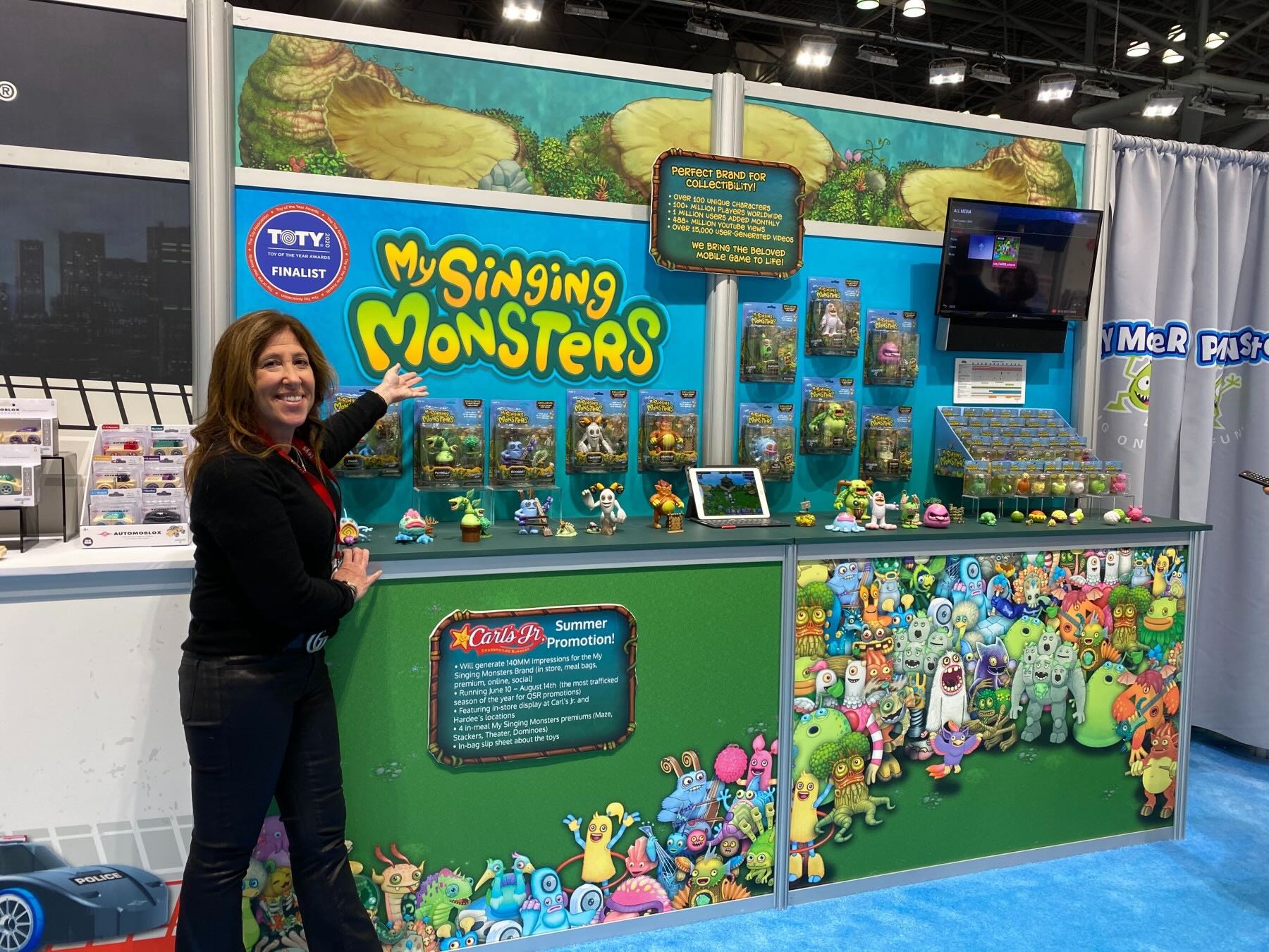 A woman stands beside a colorful My Singing Monsters display, pointing toward the large My Singing Monsters sign. The booth features character toys, boxed figures, promotional signs, a game screen, and artwork of colourful monsters.