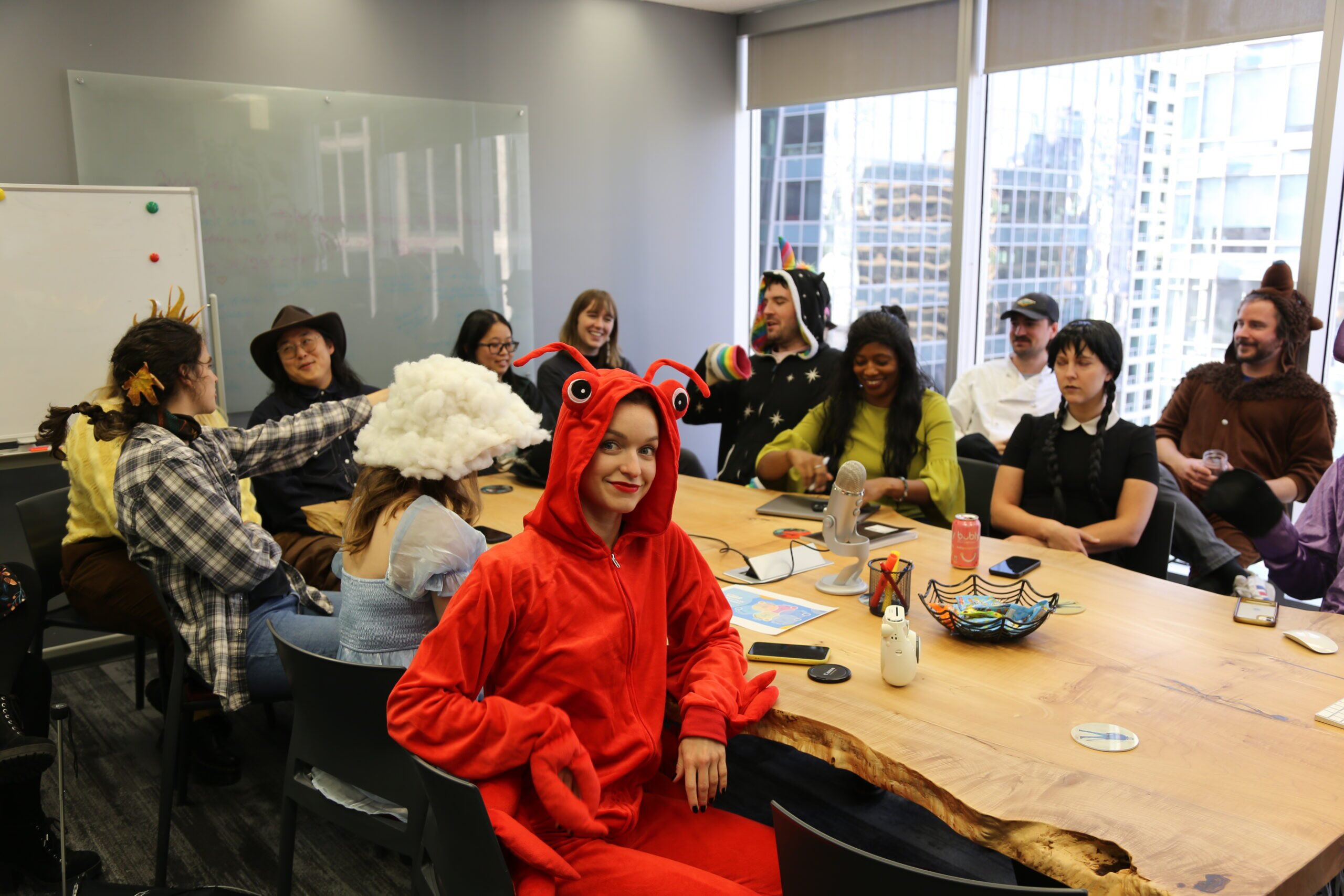 A group of coworkers in Halloween costumes gather around a large wooden conference table, with a woman in a red lobster costume smiling at the camera in the foreground. Around the table, others wear costumes including a cowboy hat, a cloud-shaped headpiece, a unicorn, a bear, and a Wednesday Addams outfit. Also the image shows floor-to-ceiling windows showing city buildings in the background.