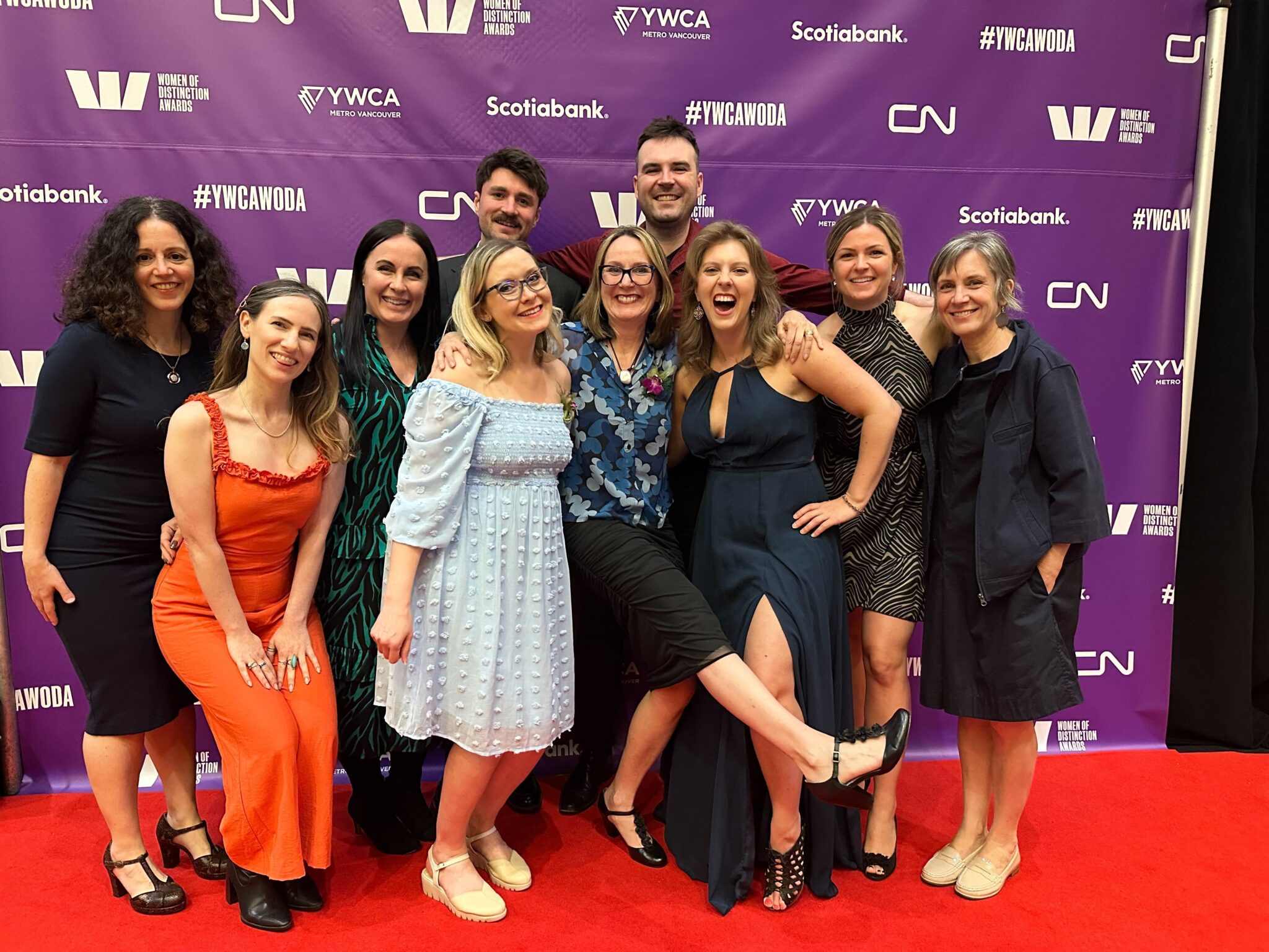 A group of nine people in dressy outfits pose together on a red carpet at the YWCA Metro Vancouver Women of Distinction Awards, smiling and laughing, with one woman playfully kicking up her leg in the front. Behind them is a purple backdrop featuring logos for the Women of Distinction Awards, YWCA Metro Vancouver, Scotiabank, and CN. Featuring Catherine Winder, CEO of Wind Sun Sky, and members of the team.